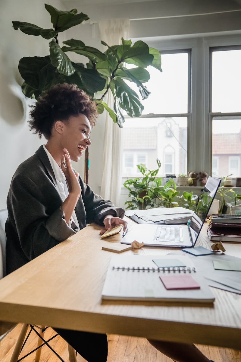 Smiling Woman Sitting in an Office and Waving to a Laptop Screen