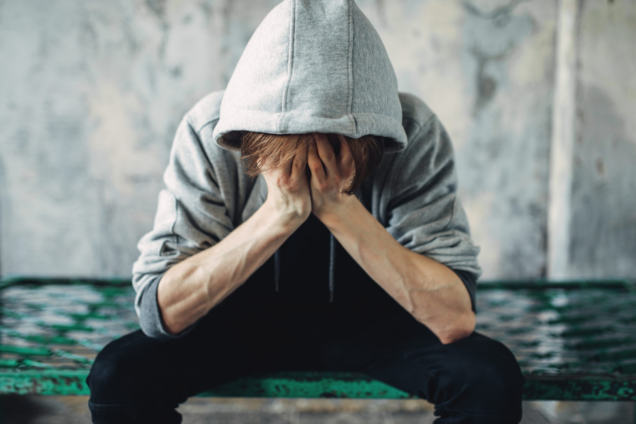 a young man sitting on the edge of his bed in a hoodie with his head buried in his hands. He is suffering because he quit drinking cold turkey