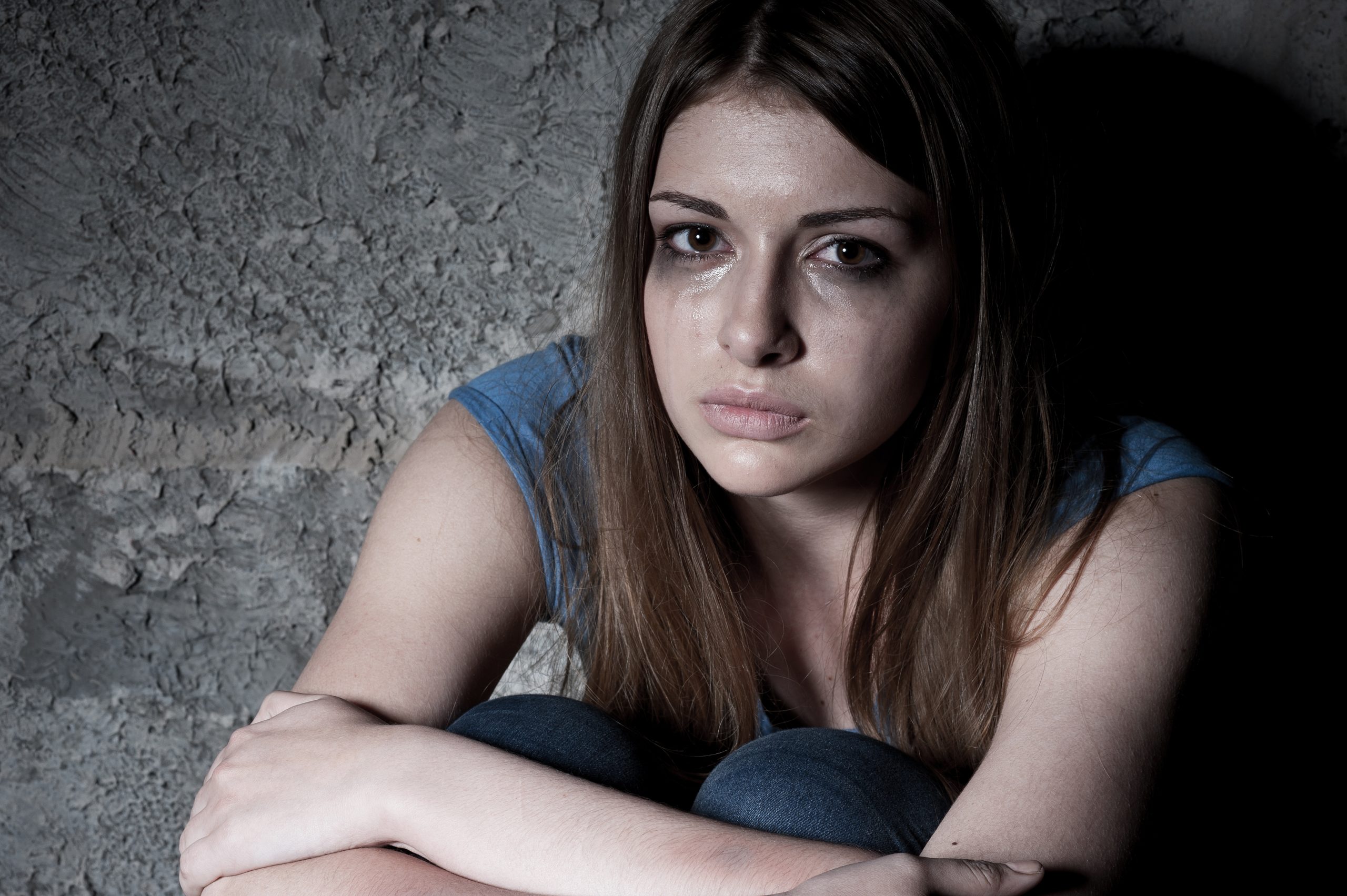 A view of a meth addict, who is a young woman, crying at looking at us while sitting against a wall